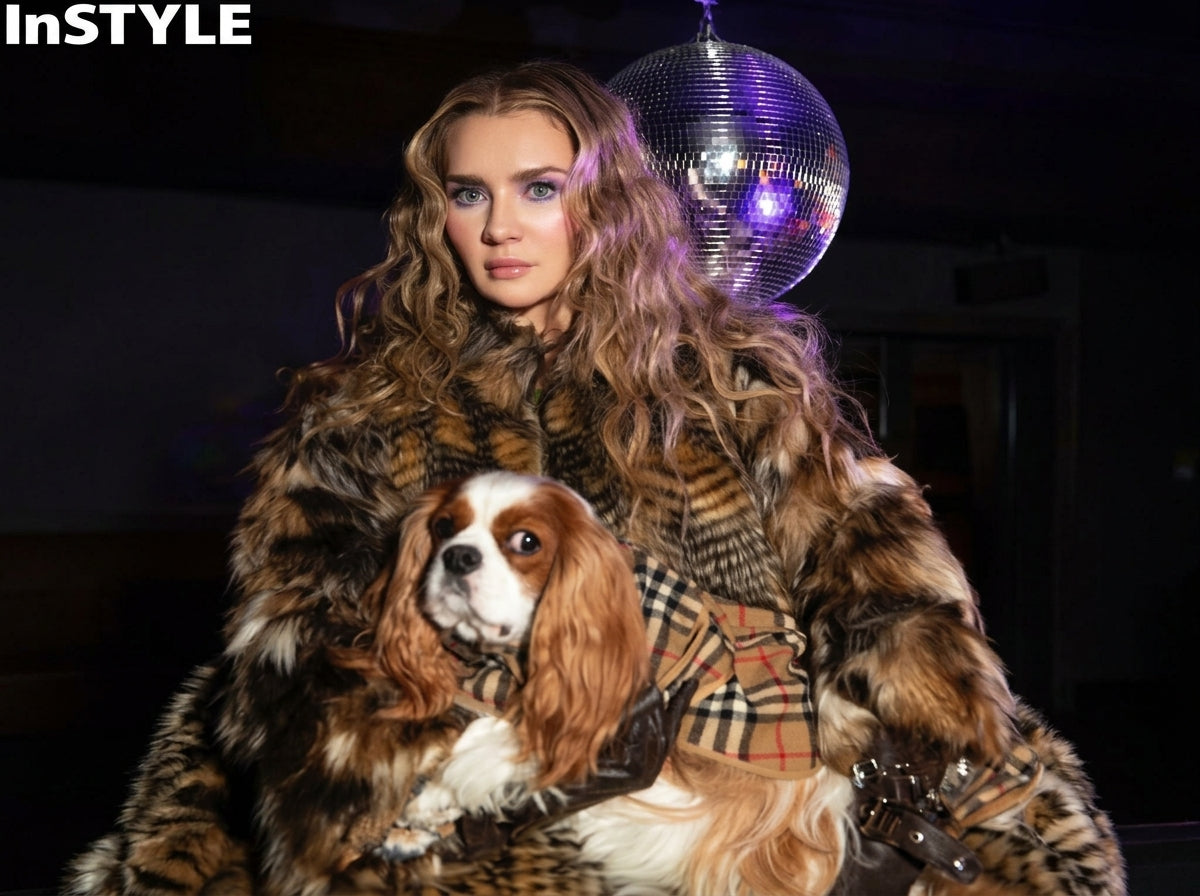 A woman in a brown and black animal-print faux fur coat holds a Cavalier King Charles Spaniel dog wearing a brown and white check harness. She is posed in front of a silver disco ball with the InStyle logo in the top left corner.