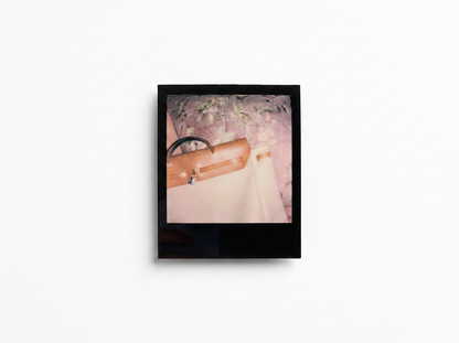A square black-framed art print or photograph hanging on a white wall, showing a soft-focus pink and beige scene with a tan handbag and pale fabric.