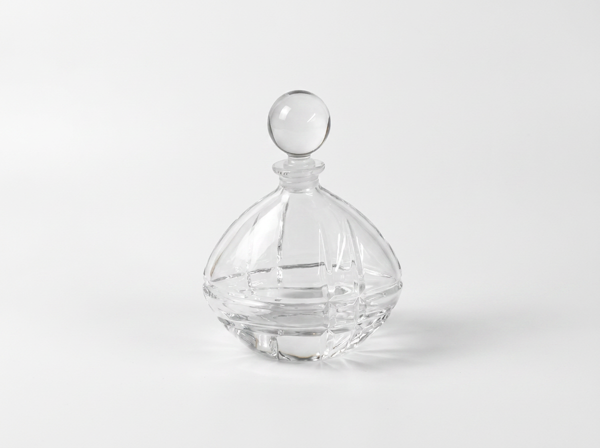 A clear glass decanter with a faceted diamond-patterned body and a spherical glass stopper, set against a seamless light gray background.