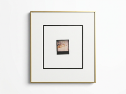 A square framed art print with a white mat, a thin gold outer frame, and a black inner border surrounding a small central photograph, shown against a plain white wall.