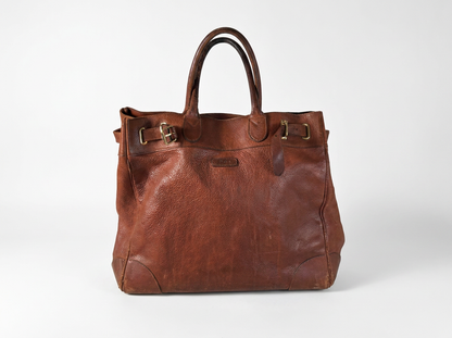 A brown leather tote bag with double top handles, brass-tone buckle details, and a small embossed brand plaque on the front, photographed against a clean white studio background.